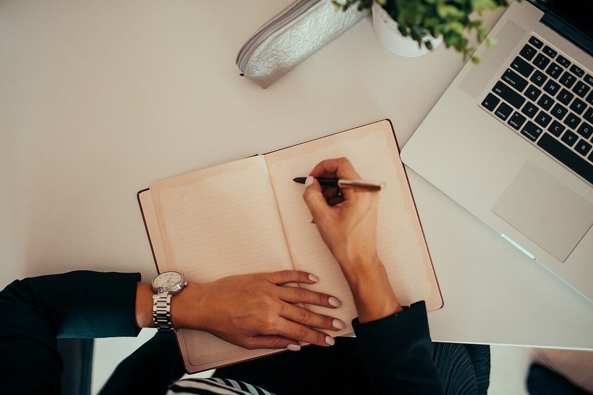 Iemand schrijft met een pen in een open notitieboek naast een laptop